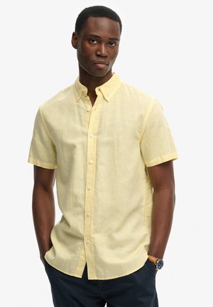 Hombre con una camisa de manga corta de color amarillo pálido con cuello y pantalones oscuros, con las manos en los bolsillos, de pie contra un fondo liso.