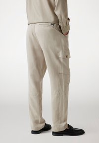 Beige cargobroek gemaakt van een lichte stof heeft twee zijzakken en zichtbare stiksels, gecombineerd met zwarte instap schoenen.