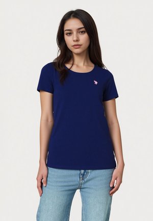 Jeune femme debout portant une chemise bleu marine à manches courtes avec un petit logo rose et un jean en denim bleu clair.