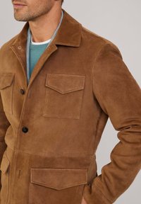 Chaqueta de ante marrón con un acabado texturizado, dos bolsillos en el pecho, cierre de botones y un cuello superpuesto sobre una camisa verde azulado.