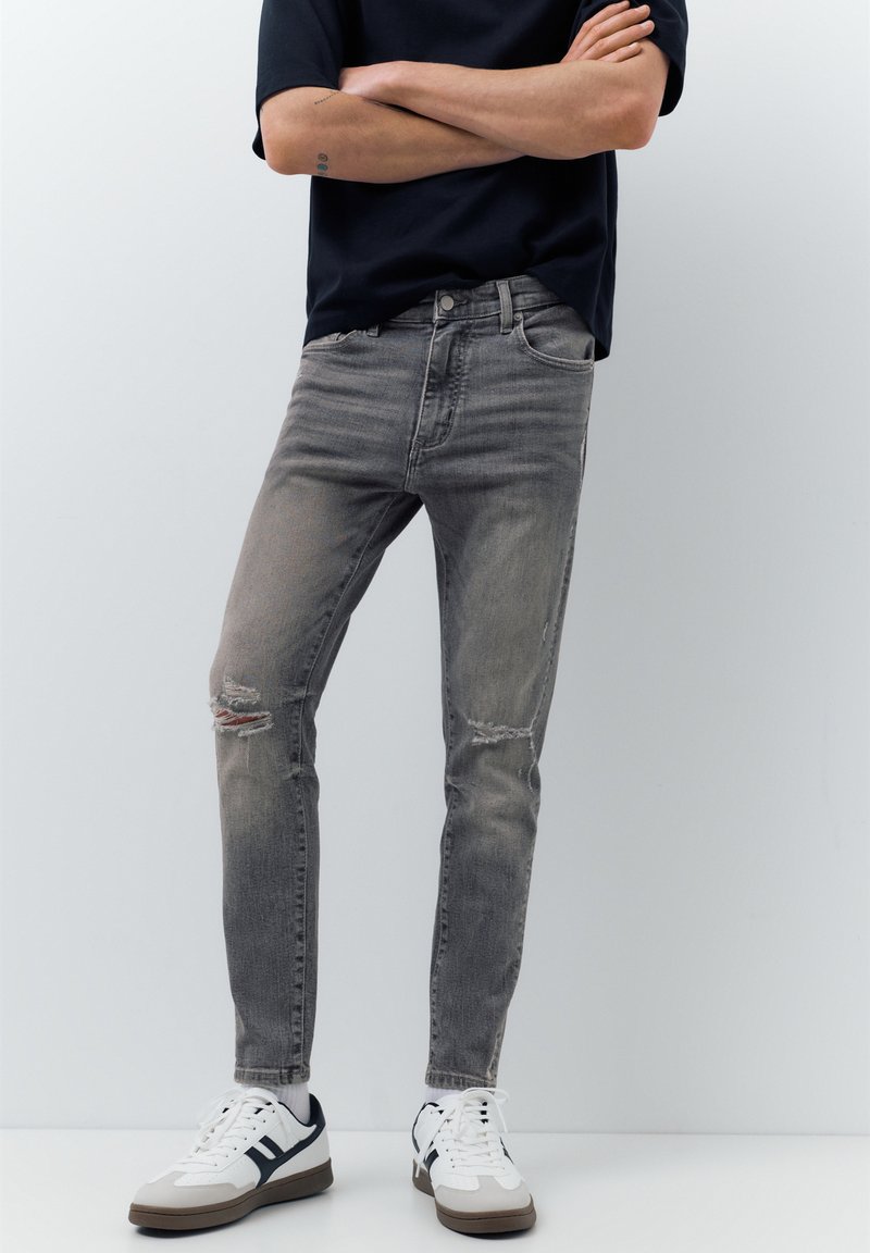 Personne portant un jean skinny gris délavé déchiré, des baskets blanches à rayures noires et un t-shirt noir à manches courtes, debout les bras croisés.