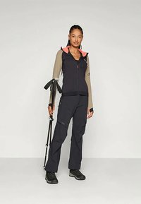 Giacca da esterno con zip di colore blu navy, maniche beige e dettagli collo rosa, abbinata a pantaloni coordinati e scarponi da trekking neri, mentre tiene in mano dei bastoncini da trekking.