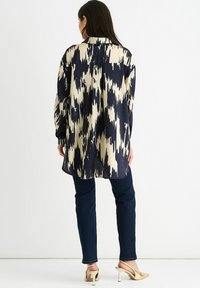 Long-sleeve shirt with a navy and cream zigzag pattern, relaxed fit, smooth fabric, and side slits, paired with dark jeans and heeled sandals.