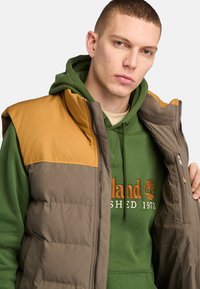 Padded vest with a brown and mustard color block design, featuring a zip closure and high collar over a green hoodie with logo details.