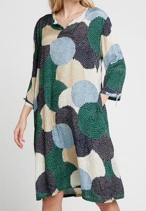 Femme portant une robe mi-longue avec de grands motifs floraux en vert, noir, bleu et beige, manches trois-quarts avec liens, et poches.