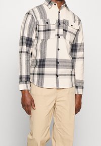 Koszula w kratę z długim rękawem w beżowych, czarnych i szarych odcieniach, z dwoma kieszeniami na piersi i czarnymi guzikami z przodu oraz na mankietach.