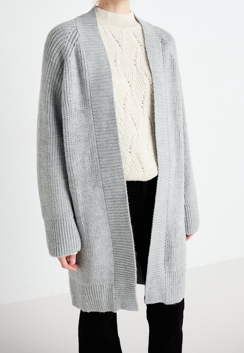 Personne portant un long cardigan gris à devant ouvert sur un pull crème à torsades et un pantalon noir, debout devant un fond blanc.