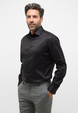 Schwarzes Langarmshirt mit schmaler Passform, glatter Textur und Knopfverschluss, kombiniert mit hellgrauen, maßgeschneiderten Hosen.