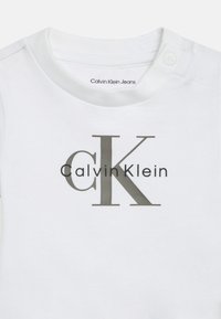 Biała bawełniana koszulka z grafiką logo w szarym kolorze, na której widnieje napis "Calvin Klein" oraz wyeksponowane inicjały "CK".