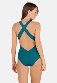 Jednodílné plavky v barvě teal s křížovými ramínky, designem ve tvaru V na zádech, vysokým pasem a hladkým povrchem. Minimalistický design neobsahuje žádné ozdoby.