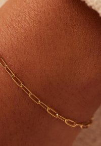 Gouden enkelband op textuurhuid, met verlengde schakels en een gladde afwerking, rustend op een warme, bruine ondergrond.
