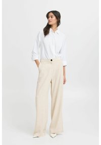 Camicia bianca con maniche arrotolate abbinata a pantaloni larghi beige. I pantaloni presentano una chiusura a un bottone e un tessuto testurizzato.