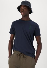 Navy blue cotton t-shirt with short sleeves, round neckline, and green accent collar. Features green embroidered number "1" and brand name.
