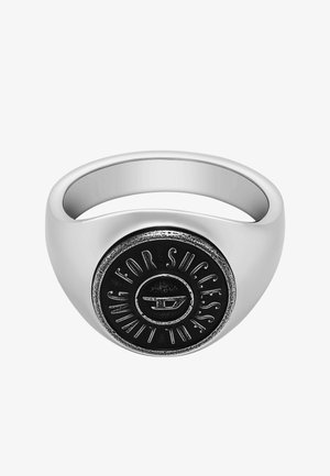 Bague en argent avec un anneau arrondi, arborant un emblème circulaire noir proéminent gravé avec "POUR LE SUCCÈS" et un design de logo au centre.
