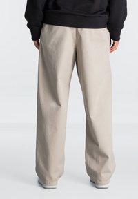 Pantalons larges beiges en coton, offrant une coupe décontractée et une texture subtile. Les ourlets du bas sont légèrement plus longs et rentrés dans des baskets blanches.