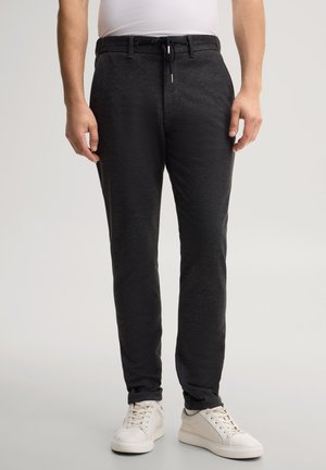 Mann trägt schwarze, strukturierte Slim-Fit-Hose mit Gürtelschlaufen, Tunnelzug in der Taille und weißen Sneakers, steht vor einem schlichten hellen Hintergrund.