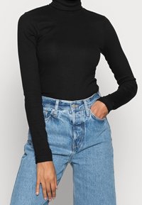 Schwarzes langärmeliges Rollkragenoberteil mit geripptem Muster, kombiniert mit hellblauen hochtaillierten Jeans im entspannten Schnitt und mit Vordertasche.