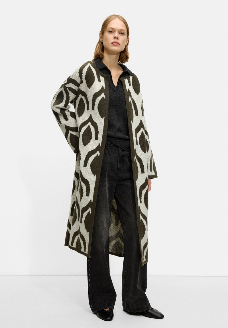 Long, open cardigan in light gray with dark green leaf patterns. Worn over a black top and black wide-leg pants, featuring a smooth texture.