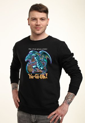 Junger Mann mit kurzem Haar und Tattoos, trägt einen schwarzen Yu-Gi-Oh!-Pullover mit einer Grafik des Blauäugigen Weißen Drachen und japanischem Text.