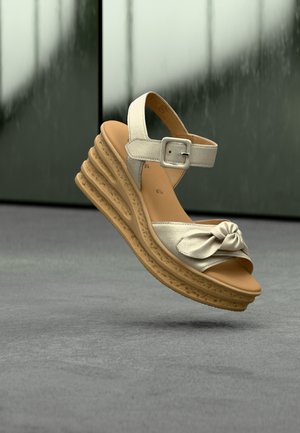 Sandale compensée beige avec design noué à l'avant, bride de cheville réglable, semelle texturée et plateforme à couches pour plus de hauteur.