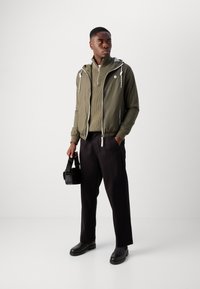 Olivgrüne Windjacke mit Kapuze, Reißverschluss vorne und weißen Akzenten; strukturierter Pullover mit hohem Kragen; schwarze Hose und Stiefel; hält eine schwarze Tasche.