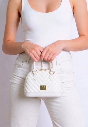 Donna con canotta bianca e pantaloni che tiene una piccola borsa trapuntata bianca con logo dorato e manici davanti a uno sfondo chiaro.