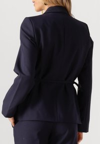 Marineblauw getailleerd colbert met een aansluitend ontwerp, lange mouwen en een ceintuur in de taille. Voorzien van een split aan de achterkant en een gladde textuur.