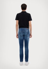 Mörk svart polo skjorta som sitter åtsittande, smala blå denimjeans med blekningar, och vita sneakers. Synliga bakfickor med dekorativ söm.