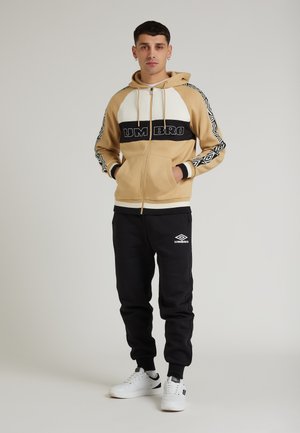 Felpa con zip beige e bianca con dettagli neri, logo "UMBRO", maniche con motivo; abbinata a pantaloni jogger neri con logo e sneakers bianche.