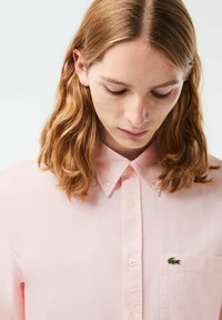 Camicia button-up di colore rosa chiaro, con colletto tradizionale e piccolo logo di un coccodrillo verde sulla tasca del petto. Tessuto di cotone liscio.