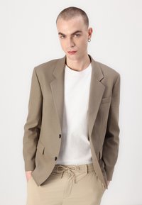 Short beige blazer with a structured design, two buttons, and a single chest pocket, worn over a white crewneck t-shirt and light beige trousers.