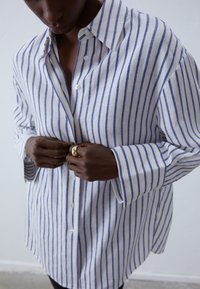 Linen shirt with blue and white vertical stripes, buttoned-up with a large collar and wide sleeves, showcasing a relaxed fit and smooth texture.