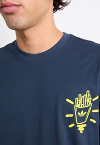 Marineblå bomulds t-shirt med et gult grafik af pomfritter i en kop med linjer strålende ud fra den. Rund hals, korte ærmer.