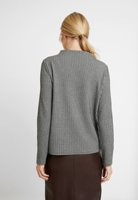 Blonde Frau mit Pferdeschwanz, die eine Jacke mit schwarz-weißen Hahnentrittmuster und einen dunkelbraunen Rock trägt, steht mit dem Rücken zur Kamera.