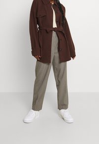 Hnědá oversized bunda s předními kapsami, zavázaná v pase, doplněná béžovou košilí a kostkovanými kalhotami. Outfit doplňují bílé tenisky.