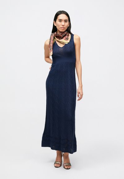 Tmavomodré pletené maxi šaty s geometrickým vzorom, výstrihom do V a žebrovaným živôtikom. Kombinované s vzorovaným šálom a sandálmi na remienky.