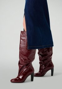 Knee-high, burgundy, patent leather boots with a slouchy design and pointed toe, featuring a thick, black stiletto heel.