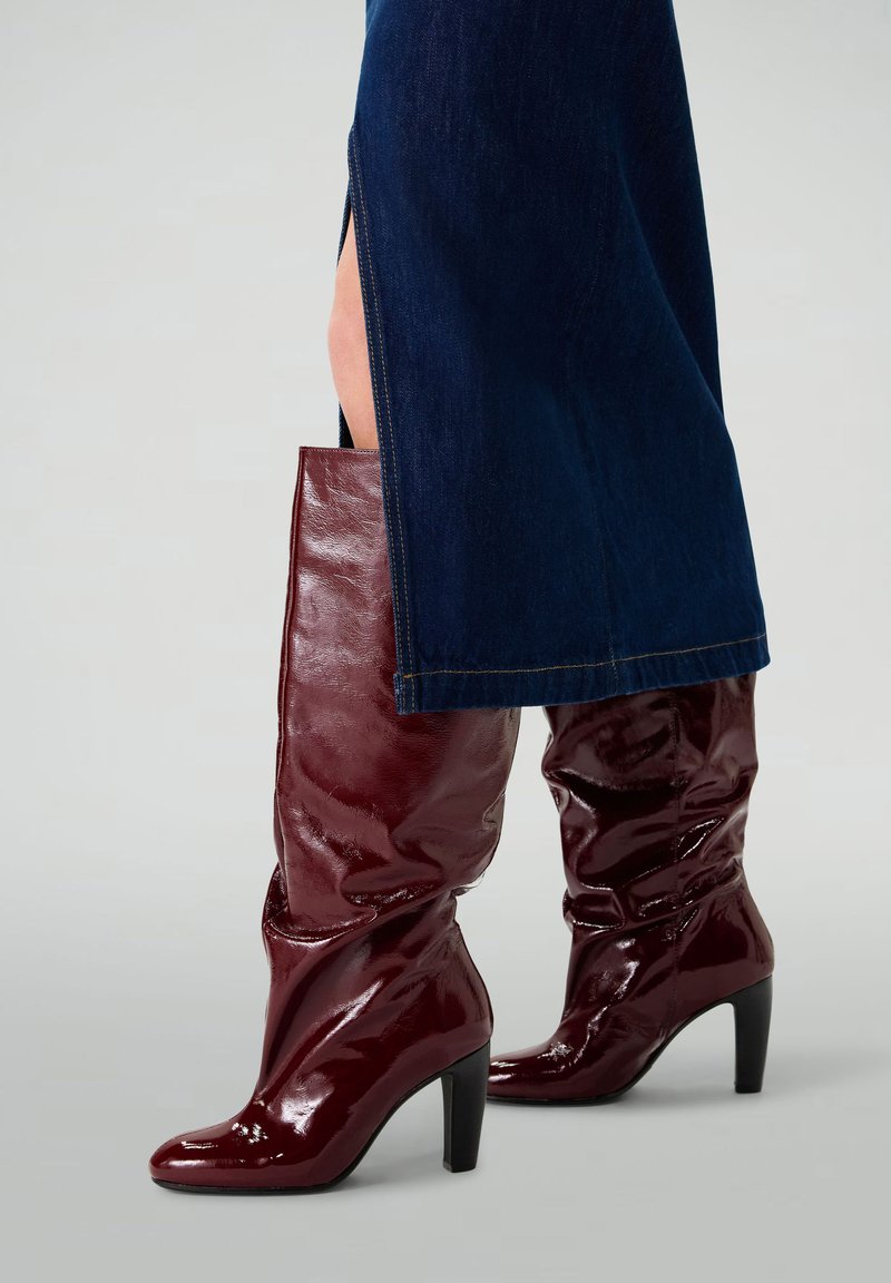 Knee-high, burgundy, patent leather boots with a slouchy design and pointed toe, featuring a thick, black stiletto heel.