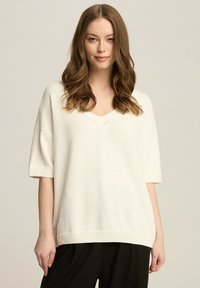 Biały sweter z dzianiny w serek, z krótkimi rękawami, o luźnym kroju i prążkowanym wykończeniu. Tekstura wydaje się miękka i lekka.