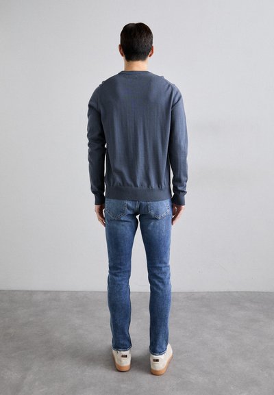 Blue long-sleeve sweater with a rounded neckline, ribbed cuffs and hem, paired with slim-fit light blue jeans and beige footwear.