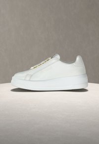 Baskets basses blanches avec semelle épaisse, œillets dorés, discrète inscription "Love Moschino" sur le côté, présentées sur une surface beige.