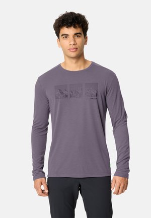 Giovane uomo in piedi che indossa una camicia a maniche lunghe viola con disegni di montagne e pantaloni neri su uno sfondo semplice.