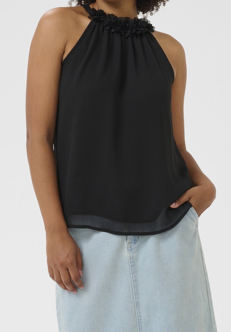 Femme portant un haut noir sans manches avec un détail floral au niveau du col et une jupe en jean bleu clair, une main dans la poche, sur un fond uni.