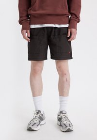 Černé cargo šortky se dvěma bočními kapsami a stahovacími šňůrkami v pase, doplněné bílými ponožkami a šedými sportovními botami. Model má na sobě vínovou mikinu.