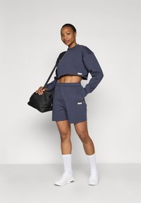 Sweatshirt cropped azul-marinho e calções combinando feitos de tecido macio. Apresenta bolsos laterais, uma mochila desportiva preta e sapatilhas brancas.