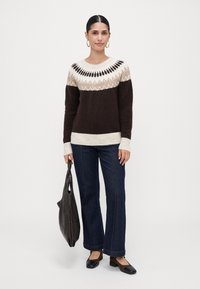 Kvinne iført en brun og beige mønstrete genser, mørkeblå jeans, svarte og brune lave hæler, og holder en svart håndveske.