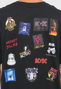 Svart bomull t-skjorte med flere fargerike AC/DC-logoer og albumomslag i forskjellige former, inkludert en lynbolt og tekst.