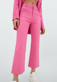 Różowe spodnie typu wide-leg wykonane z gładkiej tkaniny, z wysokim stanem, posiadające boczne kieszenie i elegancki, dopasowany krój. Połączone z białymi trampkami.