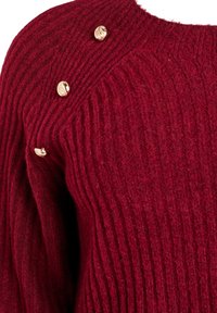 Burgunderfarbener Strickpullover mit gerippter Textur, ausgestattet mit drei metallischen Akzentknöpfen an der Schulter. Weiches, warmes Material.