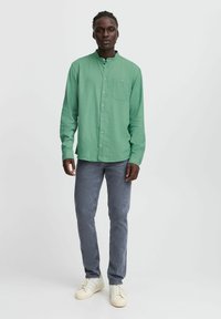 Lys grøn, langærmet skjorte med knaplukning, stående krave og en enkelt lomme, parret med grå jeans og hvide sneakers.